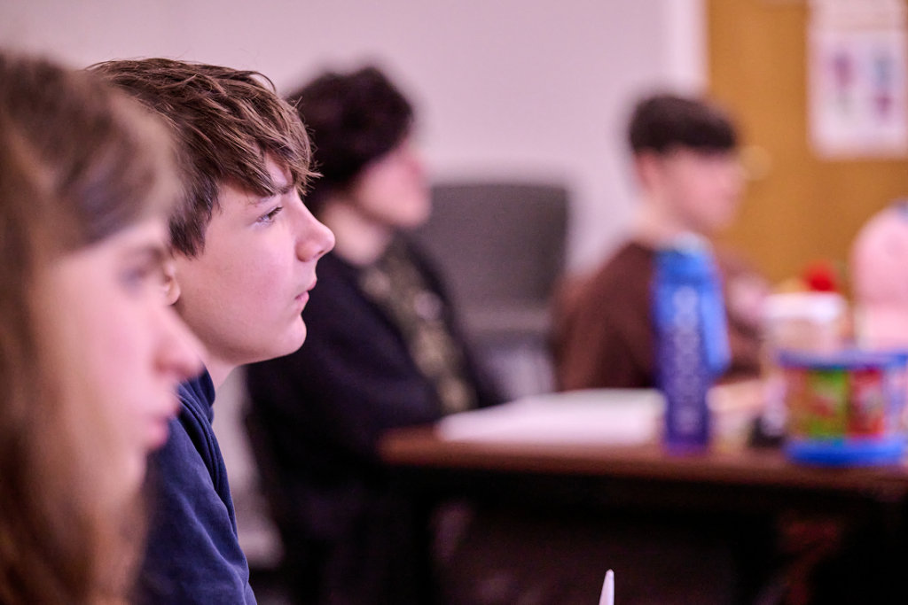 A row of pupils face the whiteboard in class, with the focus on one pupil listening attentively