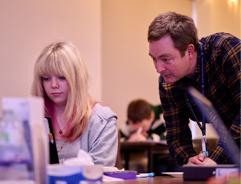 A male teacher leans over beside a blonde pupil to look at her laptop screen as she works