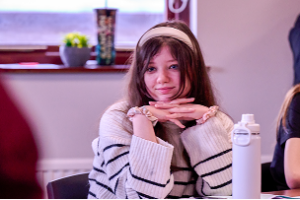 A smiling schoolgirl sits at her desk with her chin resting on her crossed fingers, looking across the classroom.